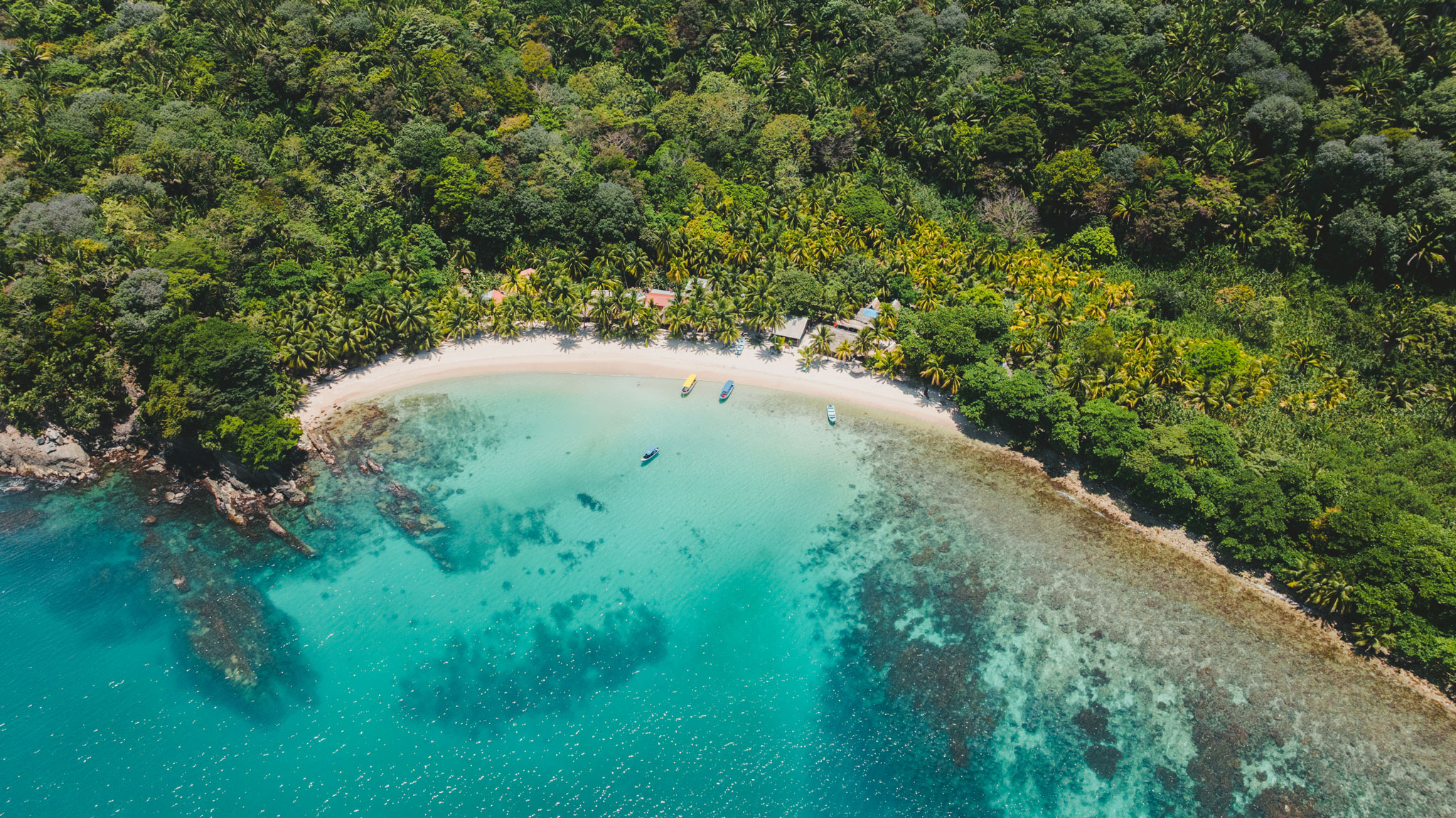 Vista aerea di una piccola e rigogliosa località costiera con una spiaggia incontaminata, acque turchesi e una barriera corallina